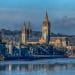 Panoramic view of Truro Cathedral in Cornwall, UK, featuring its iconic twin spires and central tower. The cathedral is set against a backdrop of lush greenery and a bright blue sky. In the foreground, traditional stone buildings line a calm river, enhancing the picturesque setting. This historic landmark is a prime example of Gothic Revival architecture in the heart of Truro. - Mike Martin Window Systems